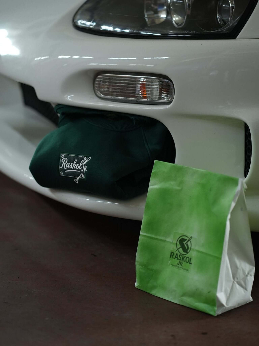 Raskol JR. Nefti Yeşil Sweatshirt displayed near a white car with Raskol shopping bag.